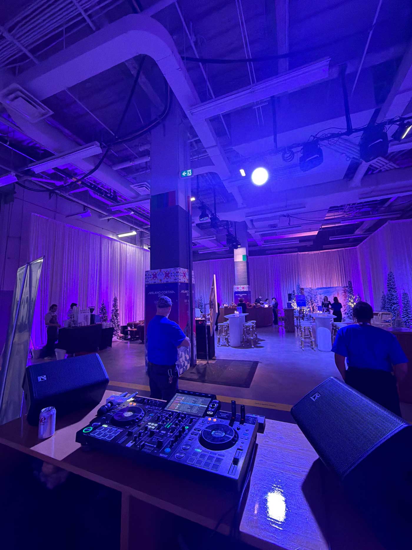 Indoor event space with purple lighting, DJ equipment and speakers on a table in the foreground, and people standing near decorated trees and tables in the background.